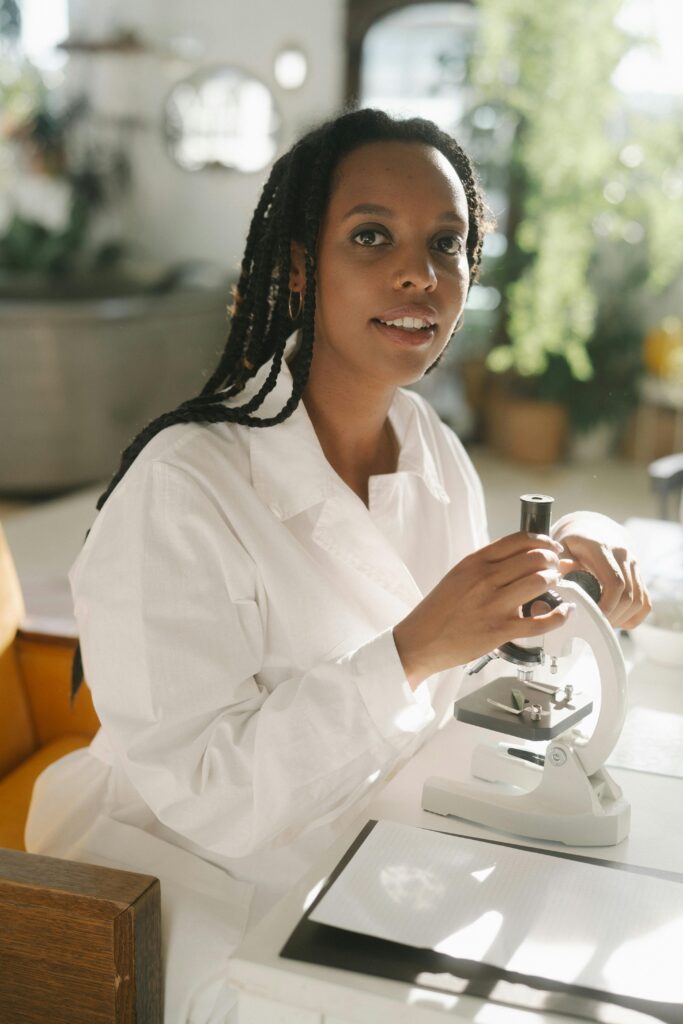 African American woman in lab gown using microscope in sunny laboratory setting.